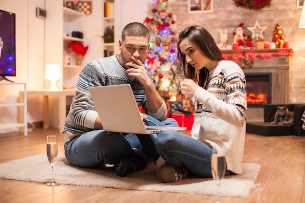 Gelukkige paar op zoek naar kerstcadeaus op internet met behulp van hun laptop voor open haard.