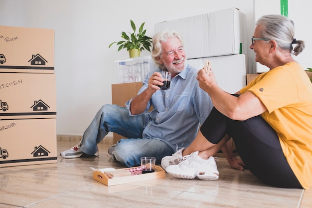 Gelukkige paar senioren die op de vloer zitten en een pauze hebben met eten en drinken in het nieuwe huis voor een nieuw begin zoals gepensioneerd met verhuisdozen op de vloer