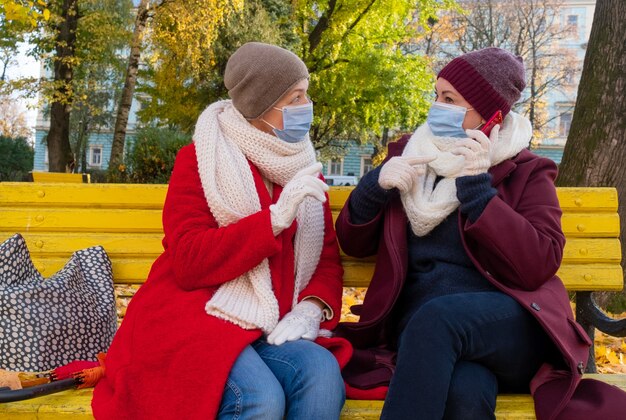 Gelukkige paar senioren of vrouwen van middelbare leeftijd dragen beschermende medische masker en zittend op een bankje in de herfst park.