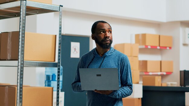 Gelukkige persoon die voorraad inventariseert met laptop in magazijn, goederen en kartonnen verpakkingen controleert voordat goederen worden verzonden. Werknemer die naar producten kijkt, voorraadbeheer. Schot uit de hand.