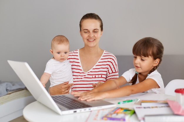 Gelukkige positieve jonge volwassen moeder met baby in handen die aan tafel zit en haar oudste dochter helpt om thuistaak te doen of te helpen tijdens de les, glimlachend op laptopcomputer.