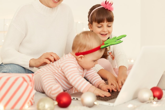 Gelukkige positieve meisjes poseren met moeder op bed tijdens oudejaarsavond voor laptopcomputer, tijd samen doorbrengen, kijken naar kersttekenfilms in de avond.