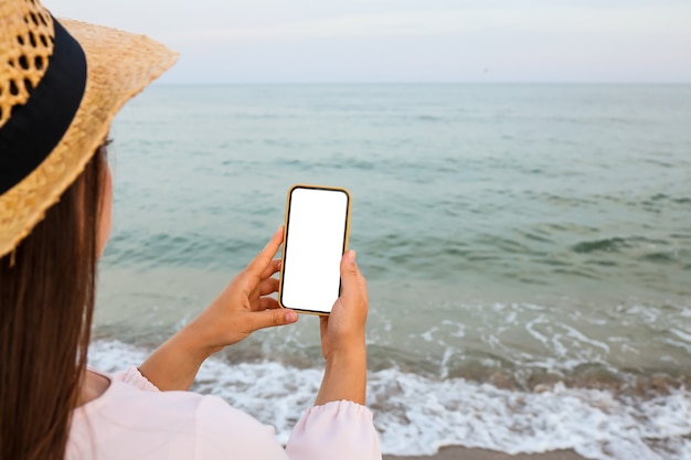 Gelukkige reiziger vrouw in roze jurk met behulp van mobiele telefoon op tropisch strand. Zomer, vakantie, technologie en lifestyle concept. Wit scherm mock-up.
