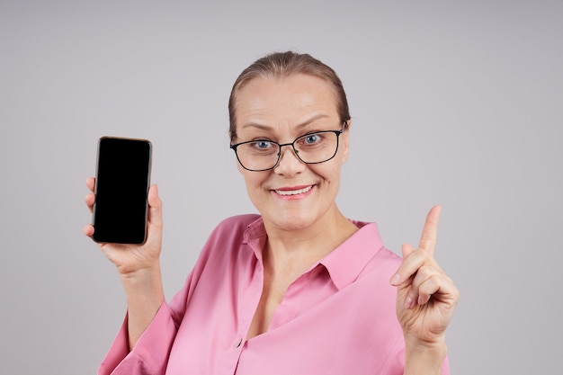 Gelukkige rijpe bedrijfsvrouw die mobiele telefoon houdt die het lege scherm toont. Geïsoleerde foto op grijze achtergrond met kopie ruimte.