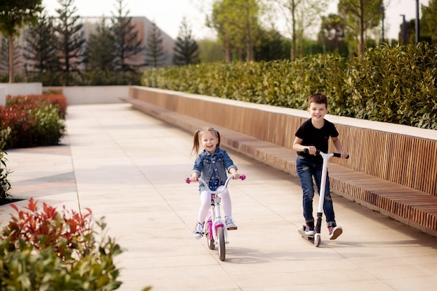 Gelukkige schattige kinderen, een jongen en een meisje fietsen en een scooter in het park in het voorjaar