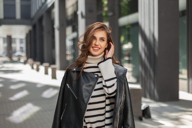 Gelukkige schattige mooie jonge vrouw met een glimlach in modieuze kleding met een leren jas en een trui loopt op een zonnige dag door de stad