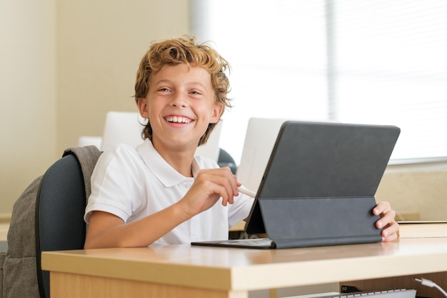 Gelukkige schooljongen met een pen op tablet tijdens de les terwijl hij aan het bureau zit in het klaslokaal op school en vooruit kijkt