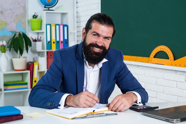 Gelukkige schoolleraar met notitieboekje die naar camera kijkt en in een klaslokaalonderwijs zit