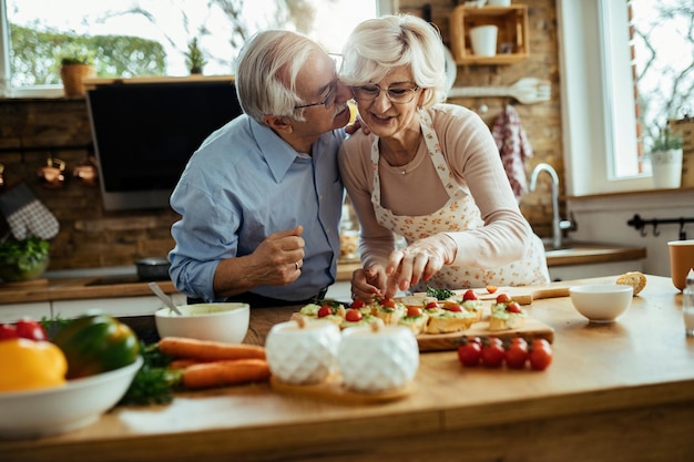 Gelukkige senior man kust zijn vrouw terwijl ze bruschetta maakt in de keuken