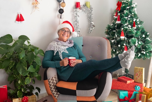 Gelukkige senior vrouw die een kerstmuts draagt na het voorbereiden van kerstcadeautjes en decoraties rust met een kopje thee - Vrolijk kerstfeest thuis voor een bejaarde gepensioneerde die op het gezin wacht