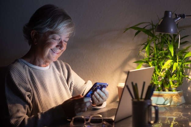 Gelukkige senior vrouw die 's avonds laat werkt en laptop gebruikt, kijkt naar het bericht van de mobiele telefoon