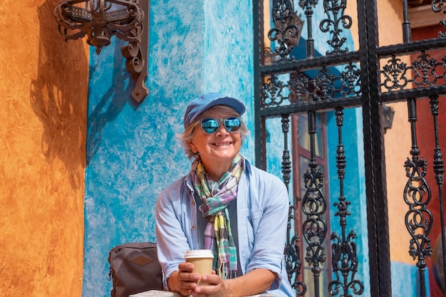 Gelukkige senior vrouw in vrijetijdsreizen zittend op een trap met een afhaalmaaltijden koffiekopje Actieve oudere dame casual kleding met pet en zonnebril