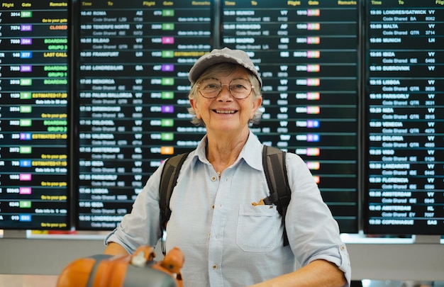 Gelukkige senior vrouw op de luchthaven die naar de camera kijkt terwijl ze naar het tijdschema kijkt om de vertrekpoort van haar vlucht te controleren Reiziger concept mensen met rugzak en koffers