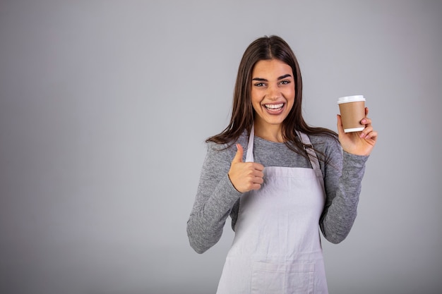 Gelukkige serveerster in schort die u glas met hete koffie aanbiedt terwijl u geïsoleerd voor de camera staat