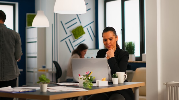 Gelukkige Spaanse dame typt op een laptop die aan het bureau zit in een startkantoor dat koffie drinkt terwijl diverse collega's op de achtergrond werken. Multi-etnische collega's die een nieuw financieel project plannen.