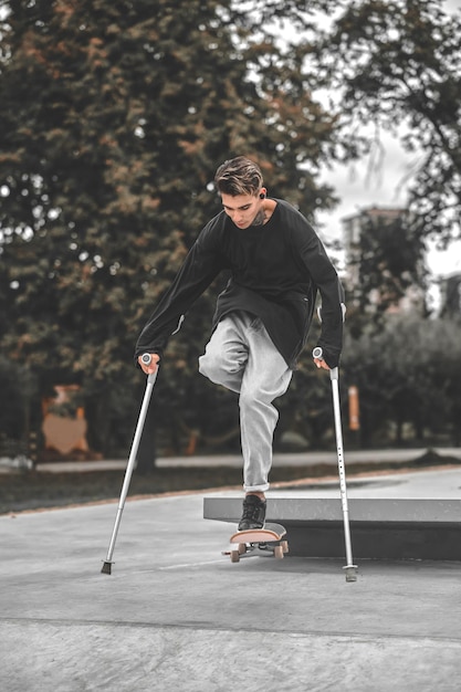 Gelukkige sprong. Betrokken schattige kerel met een handicap op krukken tijdens het springen op een skateboard vanaf de springplank in het park op een warme dag