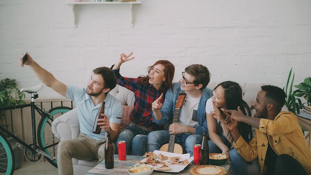 Gelukkige studentenvrienden die selfie op smartphone nemen en poseren terwijl ze feesten met bier en pizza in gedeelde accommodatie binnenshuis