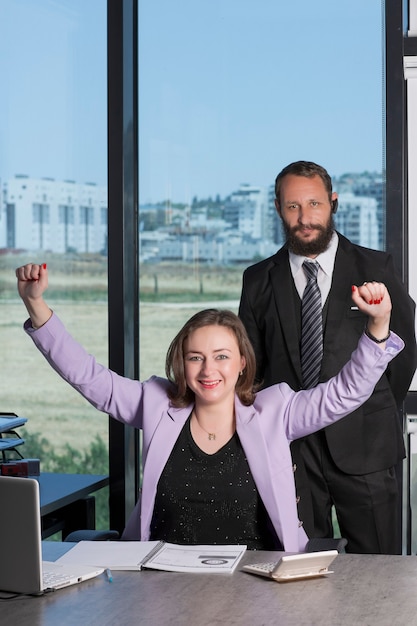 Gelukkige succesvolle glimlachende zakenvrouw stak haar handen omhoog terwijl ze aan een tafel op kantoor zat. Verenigde collega's die hun handen opsteken om hun prestatie te vieren. Succes en teamwerkconcept