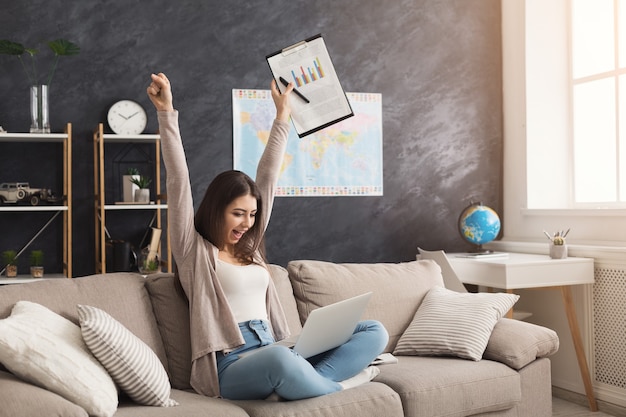 Gelukkige succesvolle vrouw die laptop gebruikt en thuis met documenten werkt terwijl ze op de bank zit. Winnaar, prestatie, succesconcept, kopieer ruimte