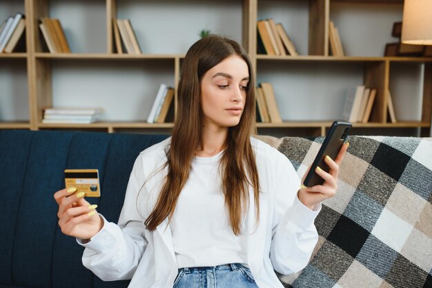 Gelukkige tiener doet online winkelen terwijl ze thuis op de bank zit Jonge vrouw voegt creditcardnummer in op de website met haar smartphone