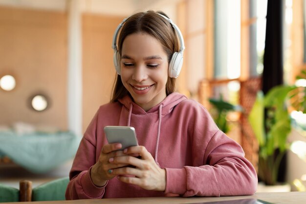 Gelukkige tiener vrouwelijke student in koptelefoon bericht aan het typen op mobiel chatten met vrienden die thuis zitten