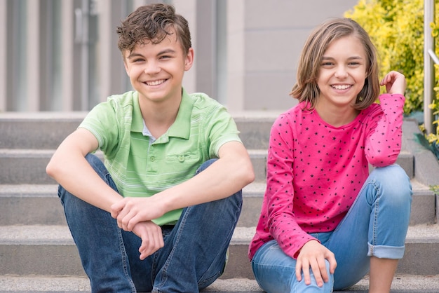 Gelukkige tienerjongen en tienermeisje glimlachen terwijl ze buiten op de trap zitten Jonge zus en broer tienerjaren kijken naar de camera