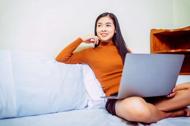 Gelukkige toevallige Aziatische vrouw die in bed met laptop werkt.