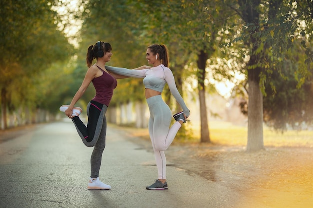 Gelukkige twee vriendinnen die sportkleding dragen, doen opwarmingsoefeningen voordat ze langs het zonnige laanpad rennen.