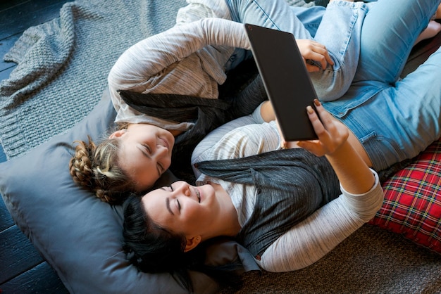 Gelukkige twee zussen die tablet gebruiken terwijl ze thuis op hun rug op de vloer liggen