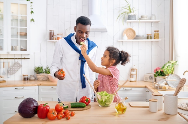 Gelukkige vader en kind koken groentesalade bij het ontbijt. Glimlachende familie eet 's ochtends in de keuken. Vader voedt vrouwelijk kind, goede relatie