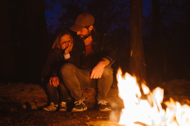 Gelukkige vader en zijn zoon warmen zich 's nachts op bij het vuur terwijl ze in een omhelzing op boomstammen zitten tijdens een wandeling in het bos. .