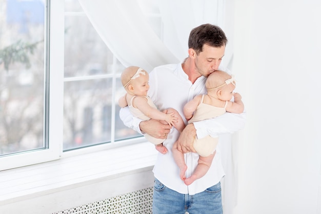 Gelukkige vader met een tweeling, papa houdt twee pasgeboren baby-tweelingmeisjes in een katoenen pak bij het raam van het huis, knuffelt en kust ze, vaderschap met twee kinderen