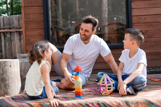 Gelukkige vader of babysitter heeft plezier met klein meisje en jongen, speelt met hen in de tuin zittend op het tapijt bij het houten huis en helpt hen kinderspeelgoed te verzamelen