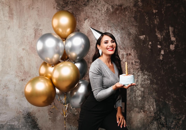 Gelukkige verjaardag Sexy brunette poseren met ballonnen vuurwerk verjaardagstaart op de achtergrond van een decoratieve muur een vrouw blaast de kaarsjes op de taart