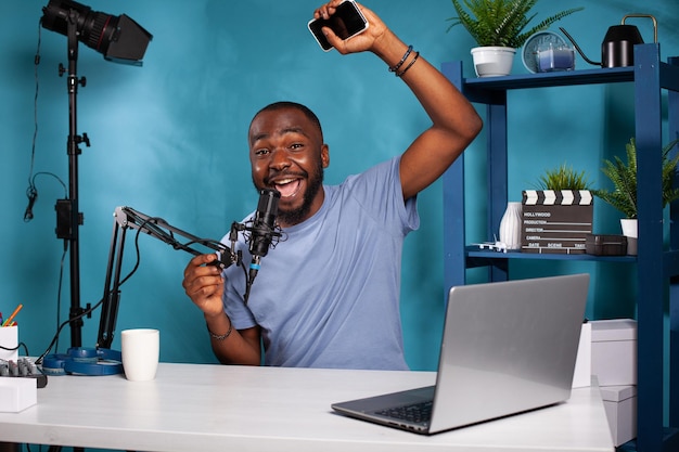 Gelukkige vlogger die hand in opwinding opsteekt terwijl hij smartphone schreeuwt in professionele microfoon die aan het bureau zit in vlogstudio. De maker van de inhoud voelt vreugde na het ontvangen van positieve feedback.