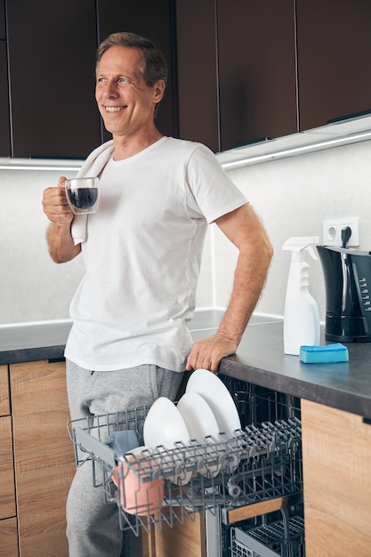 Gelukkige volwassen aardige man die glimlach op zijn gezicht houdt terwijl hij thuis geniet van koffie in de ochtend