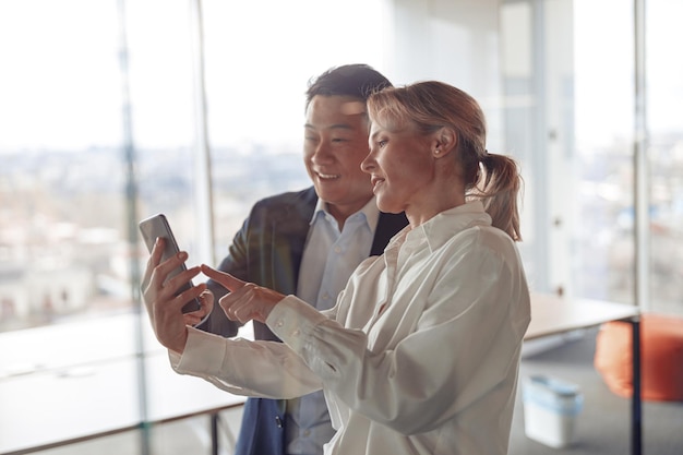 Gelukkige volwassen diverse collega's die naar smartphone kijken en glimlachen terwijl ze in de buurt van een glazen wand staan