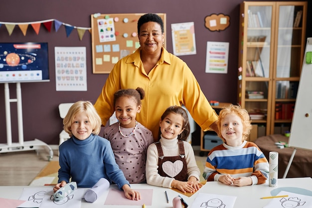 Gelukkige volwassen leraar en een groep interculturele kleine leerlingen die bij het bureau stonden en ernaar keken, kwamen