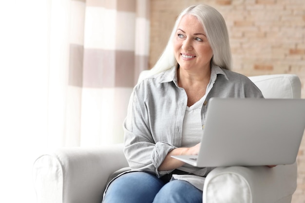 Gelukkige volwassen vrouw die laptop gebruikt terwijl ze thuis in een fauteuil zit