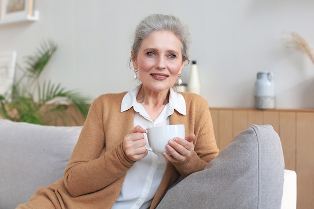 Gelukkige volwassen vrouw die op een comfortabele bank rust, drinkt koffie of thee, kijkt weg, ontspant thuis op een gezellige bank en geniet van warme drank
