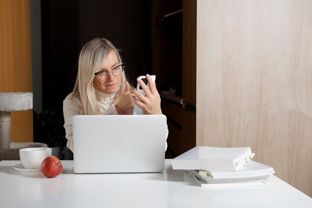 Gelukkige volwassen vrouw met laptop en smartphone werkt thuis en videochat
