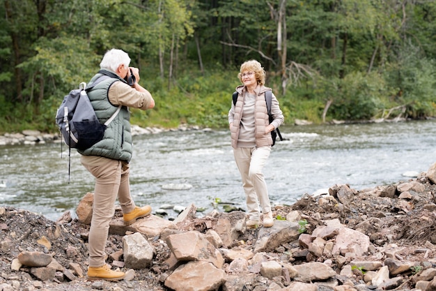 Gelukkige volwassen vrouwelijke backpacker die zich door rivier voor haar echtgenoot bevindt die met camera haar tijdens reis fotografeert