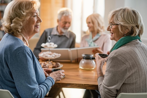 Gelukkige volwassen vrouwen die koffie drinken en thuis communiceren terwijl hun vrienden op de achtergrond naar het fotoalbum kijken