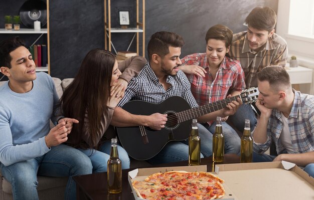 Gelukkige vrienden die gitaar spelen, bier drinken en pizza eten thuis