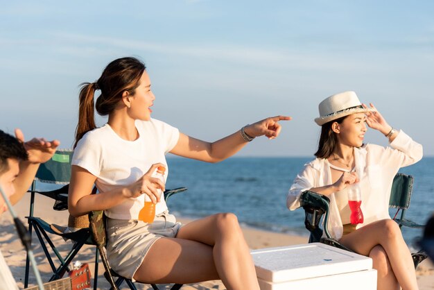 Gelukkige vrienden met drankjes in het kamp en samen lachen tijdens vakanties op zandstrand in de buurt van camping tent vakantietijd bij zonsondergang, jonge Aziatische groep vrouw en man in de zomer reizen buiten