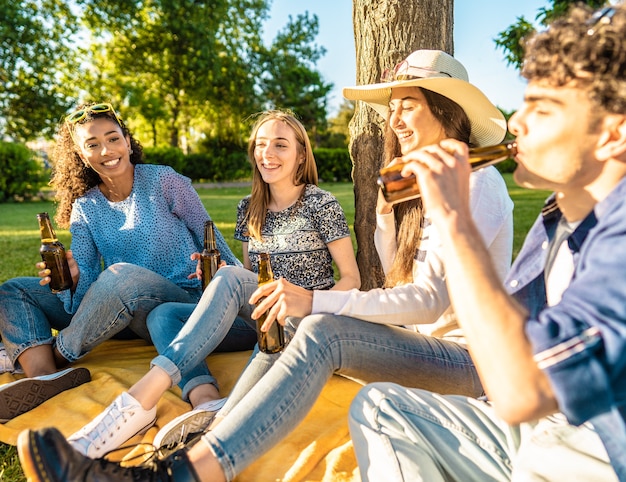 Gelukkige vriendengroep buiten in stadspark bier drinken uit fles vieren zittend op gras. Zorgeloze jonge lachende mensen die plezier hebben in de natuur met alcohol bij zonsondergang genieten van een picknick in het gras