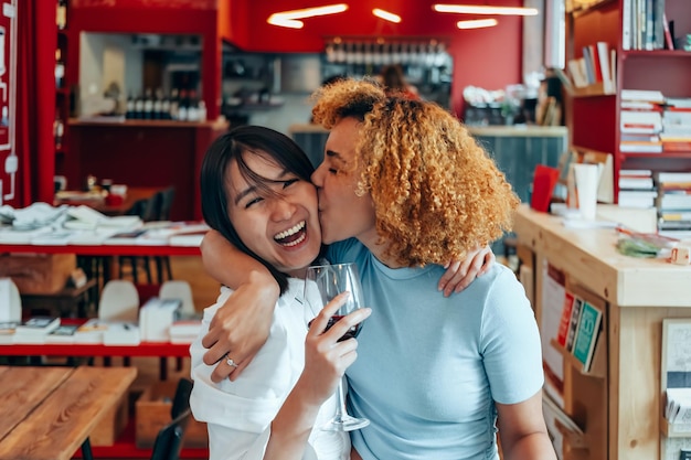 Gelukkige vriendinnen in het café omhelzen elkaar en lachen - Braziliaans meisje kust Aziatische vrouw op de wang