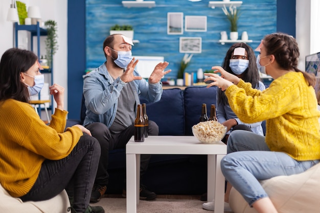 Gelukkige vrolijke multi-etnische vrienden met gezichtsmasker die spelletjes spelen met plaknotities op de voorhoofden die bier drinken en popcorn eten, in de veronderstelling dat ze sociale afstand houden. tijdens uitbraak in huis woonkamer