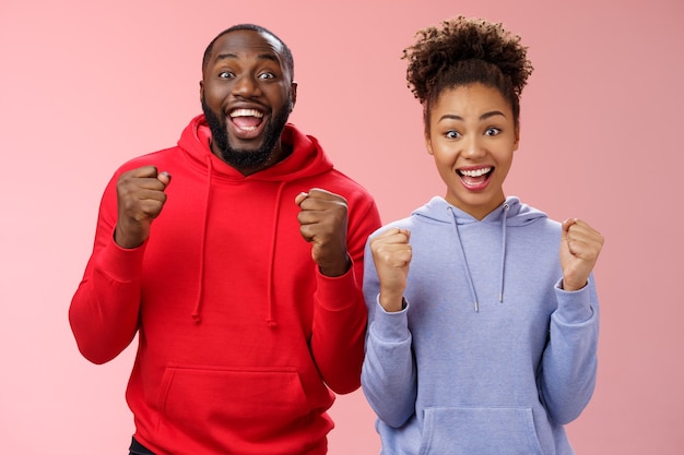 Gelukkige vrolijke twee Afro-Amerikaanse man vrouw schreeuwen hoera vieren triomfantelijk enorm succes balde vuisten vreugdevol wederzijds doel staande vreugdevol roze achtergrond overwinning gebaar