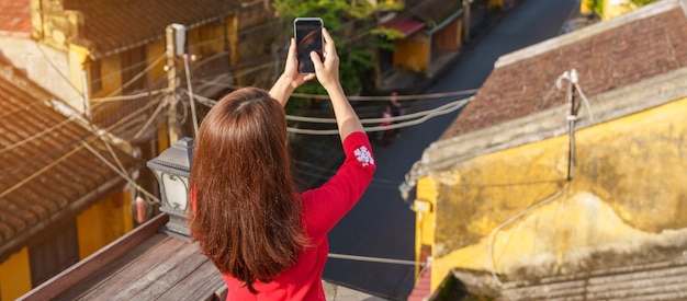 Gelukkige vrouw die Ao Dai Vietnamese kledingreiziger draagt die foto neemt door mobiele telefoon op dak bij de oude stad Hoi An in Vietnam Vietnam en Zuidoost-reisconcept
